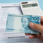Reforma Tributária: O Impacto de 5% no seu Bolso e Empresas Mão segurando cédula de Real com calculadora e documentos fiscais ao fundo, simbolizando o impacto da reforma tributária no Brasil.
